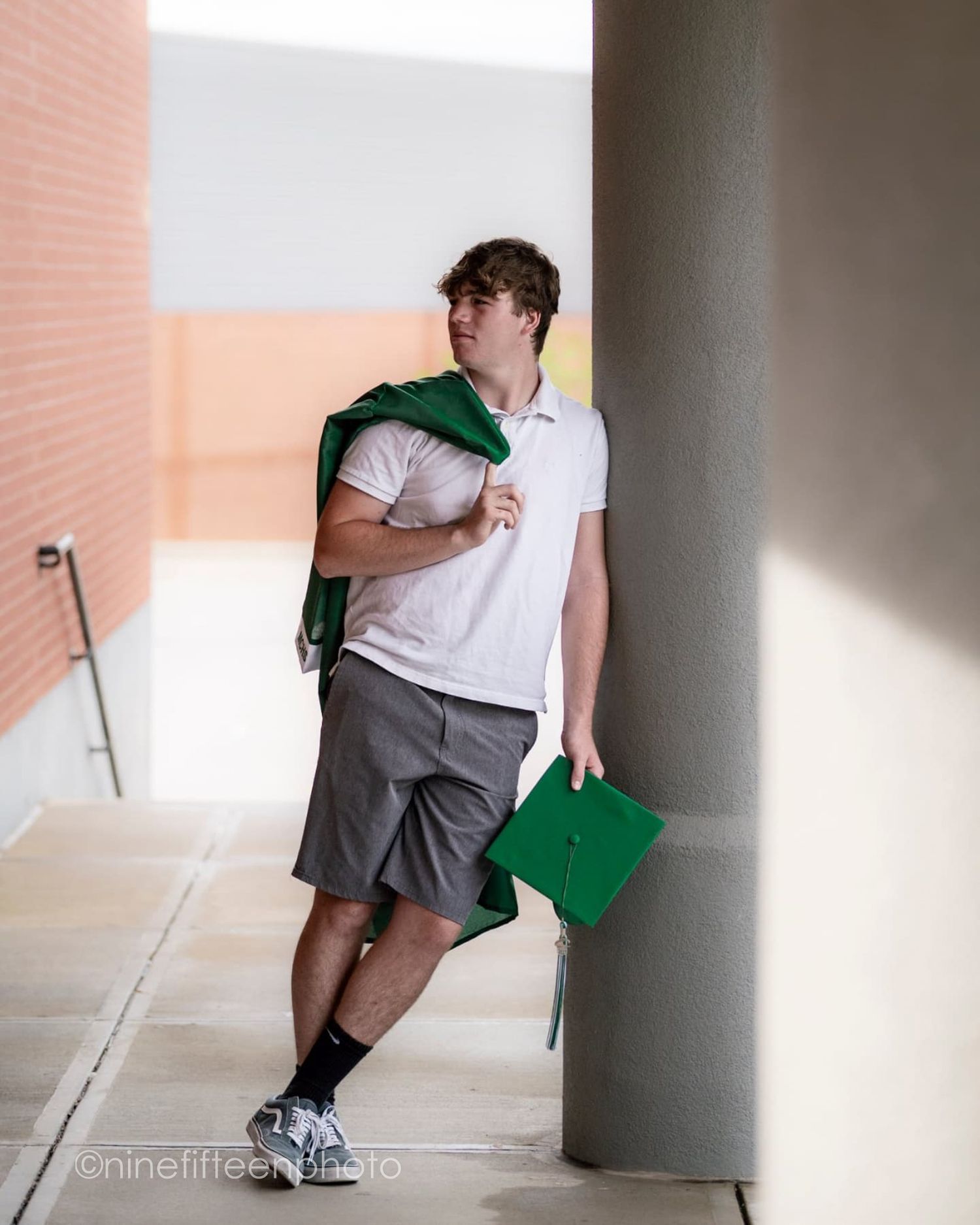 Young man in white polo and gray shorts holding green graduation cap and gown, leaning against a column.