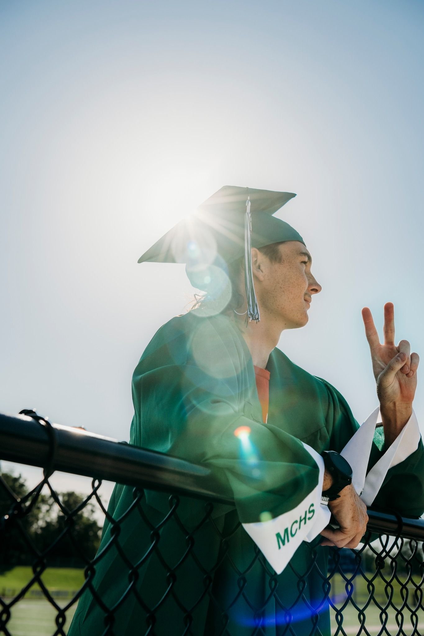 Male graduate in green MCHS cap and gown flashes a peace sign in bright sunlight.