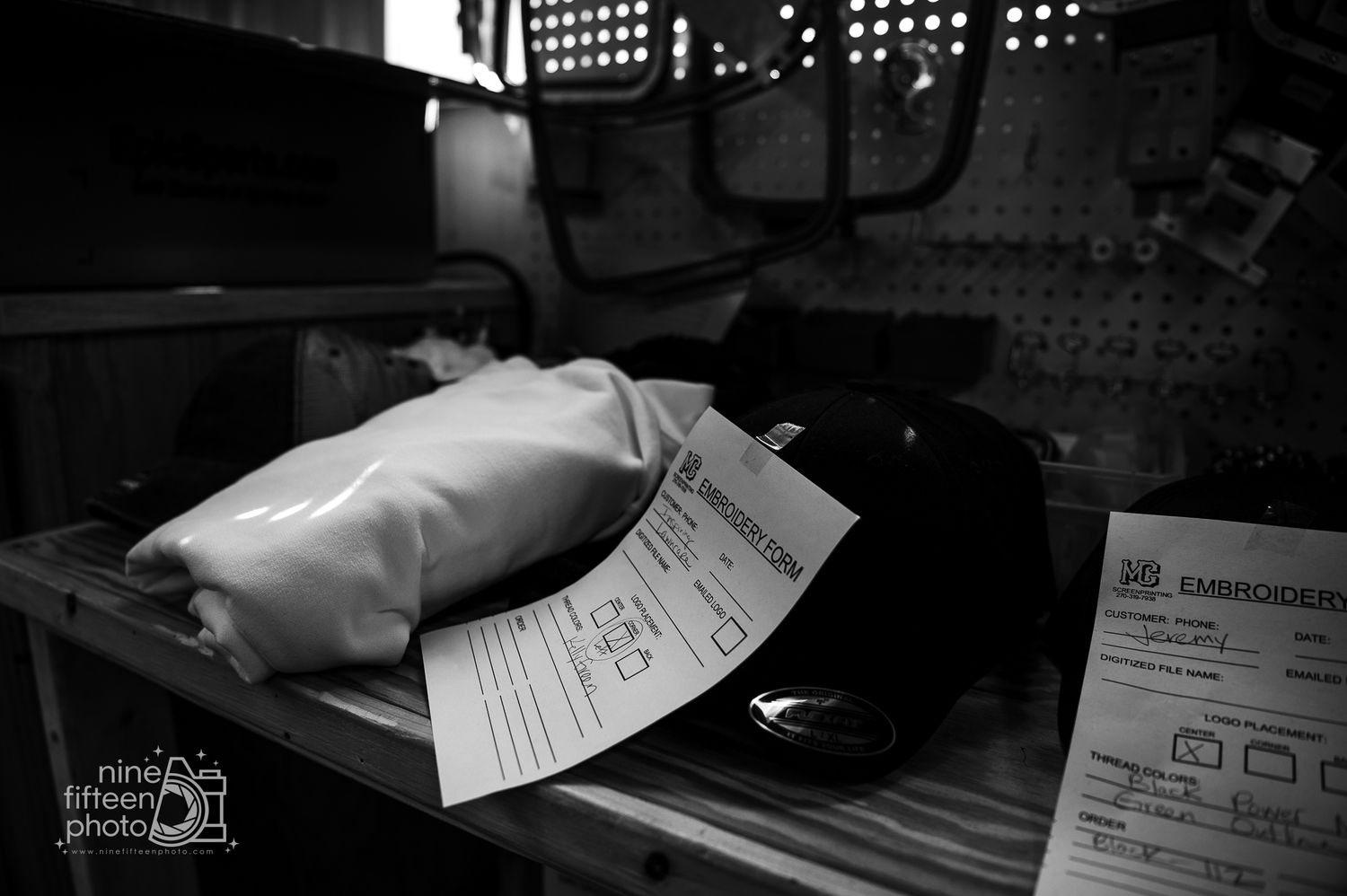 Black and white photo of embroidery order forms and garments on a workbench.