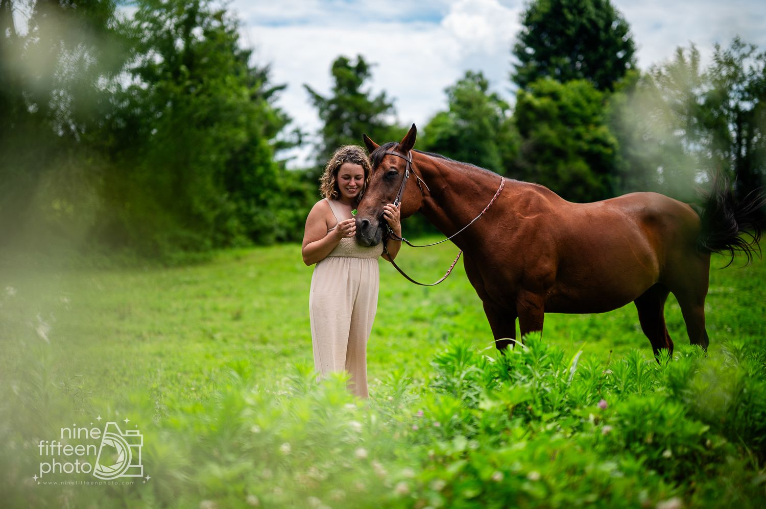 Woman in white dress standing with a chestnut horse in a lush green field, professional equine photography.
