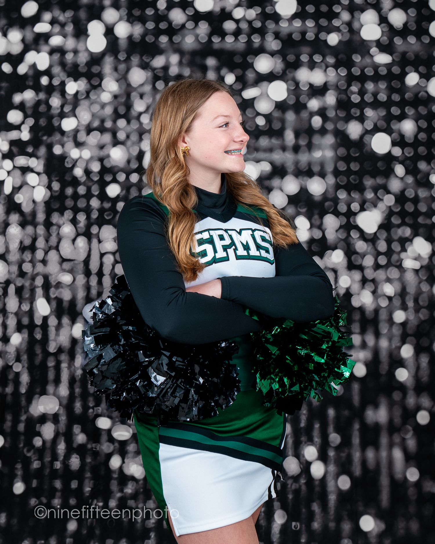 A cheerful teenage girl in a SPMS green and white cheerleading uniform poses with pom-poms against a silver sequin backdrop.