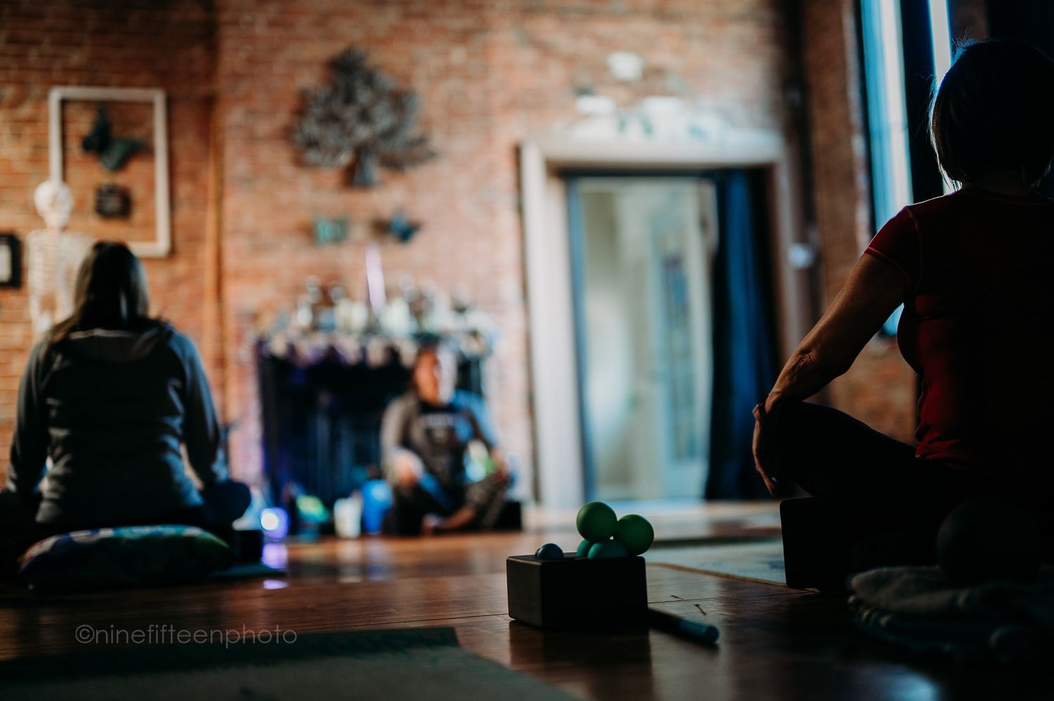 Two people sitting in silent meditation on yoga mats in a dimly lit brick studio.