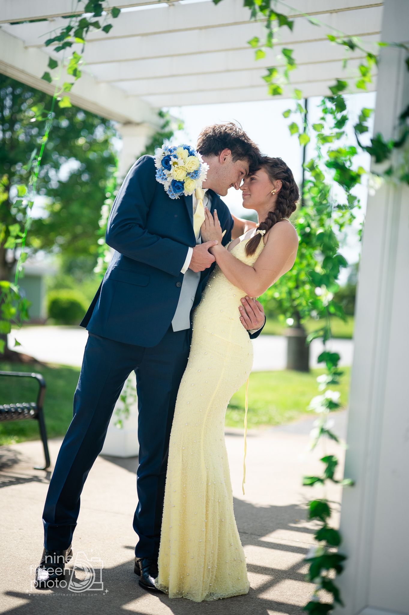 Couple in formal wear sharing a romantic moment under a floral arch, man in navy suit, woman in yellow gown.