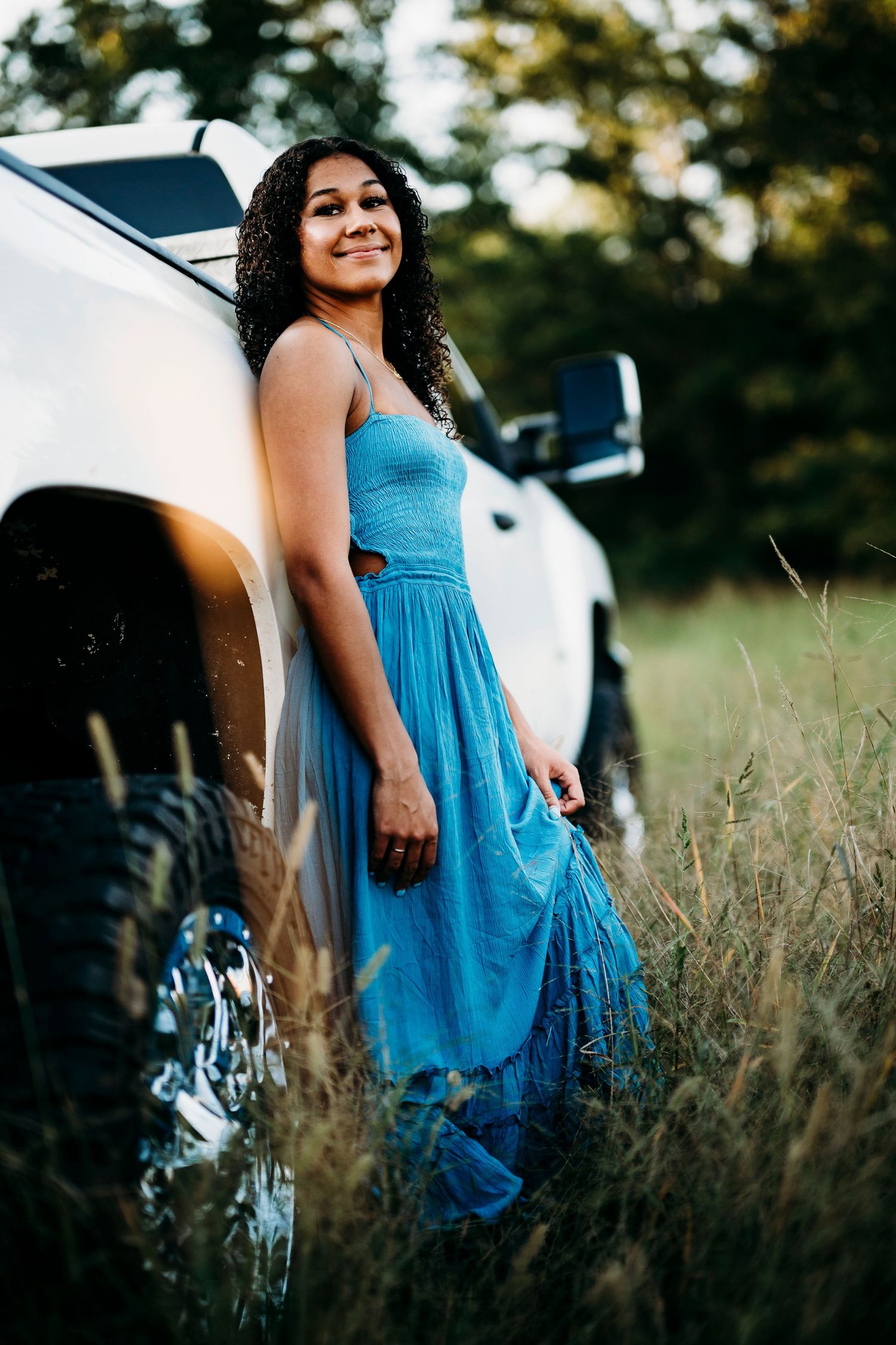 Senior in bright blue dress smiling outdoors surrounded by tall grass at sunset.