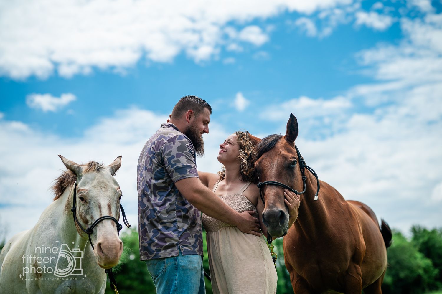 Couple in wedding attire stands between a white and brown horse under a blue sky in an outdoor engagement photo.