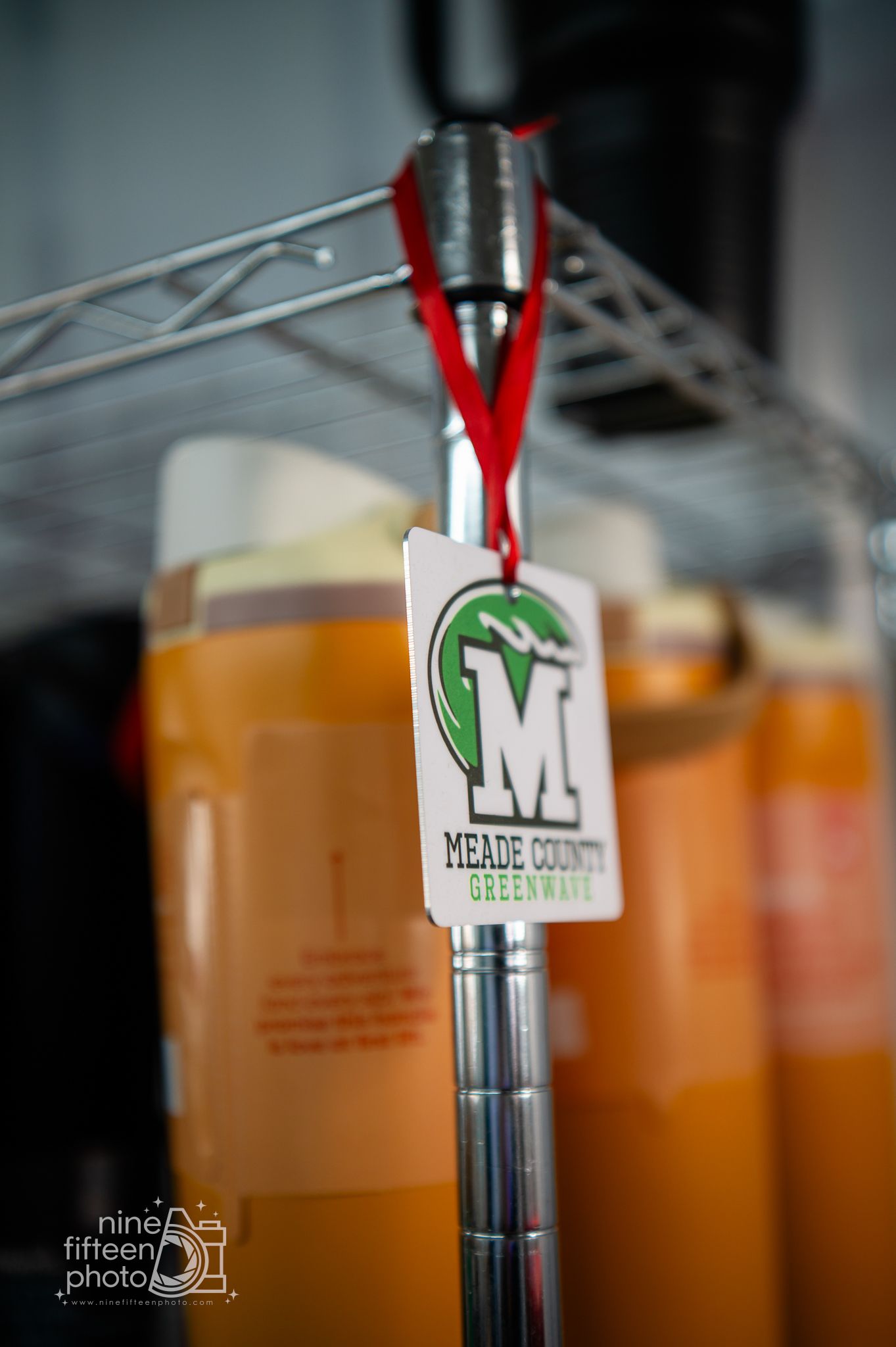 Meade County Greenwave logo tag hanging on a metal shelf rack with orange liquid containers.