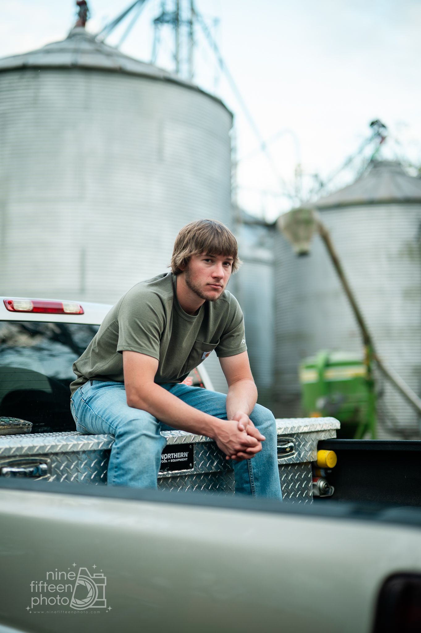 Young man in green t-shirt and jeans sitting on a truck tailgate with grain silos in the background.
