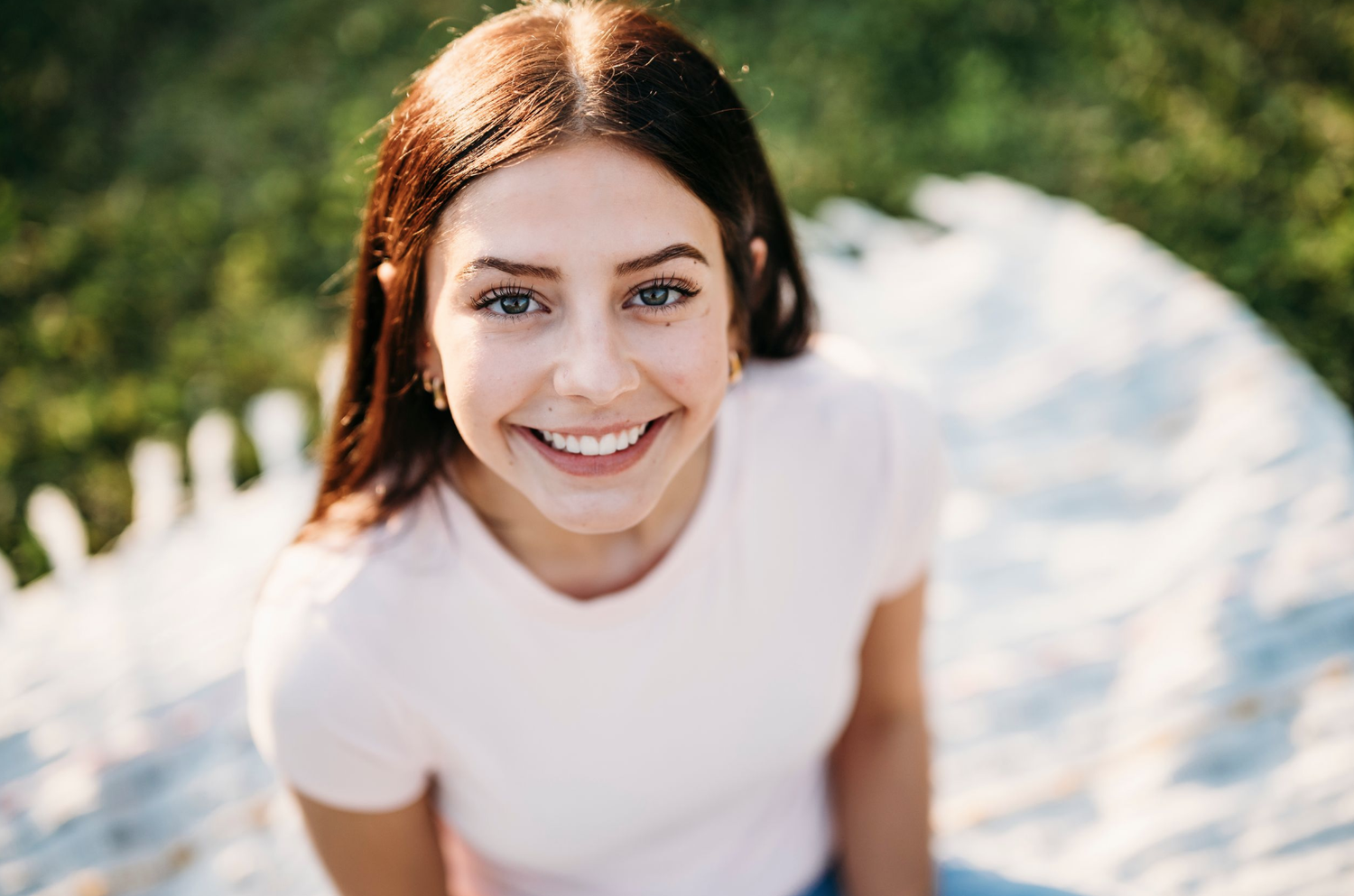 Smiling young woman with dark hair in a white t-shirt looking up at the camera outdoors in sunlight.