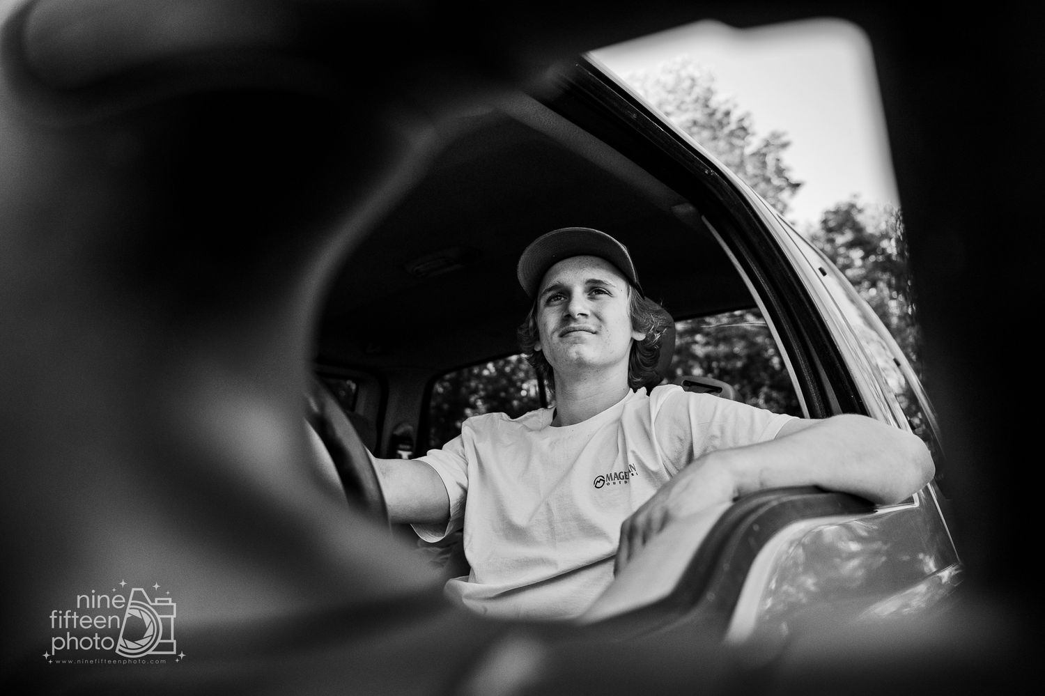 Black and white photo of a young man in a cap sitting in a car, shot creatively through a side mirror reflection.