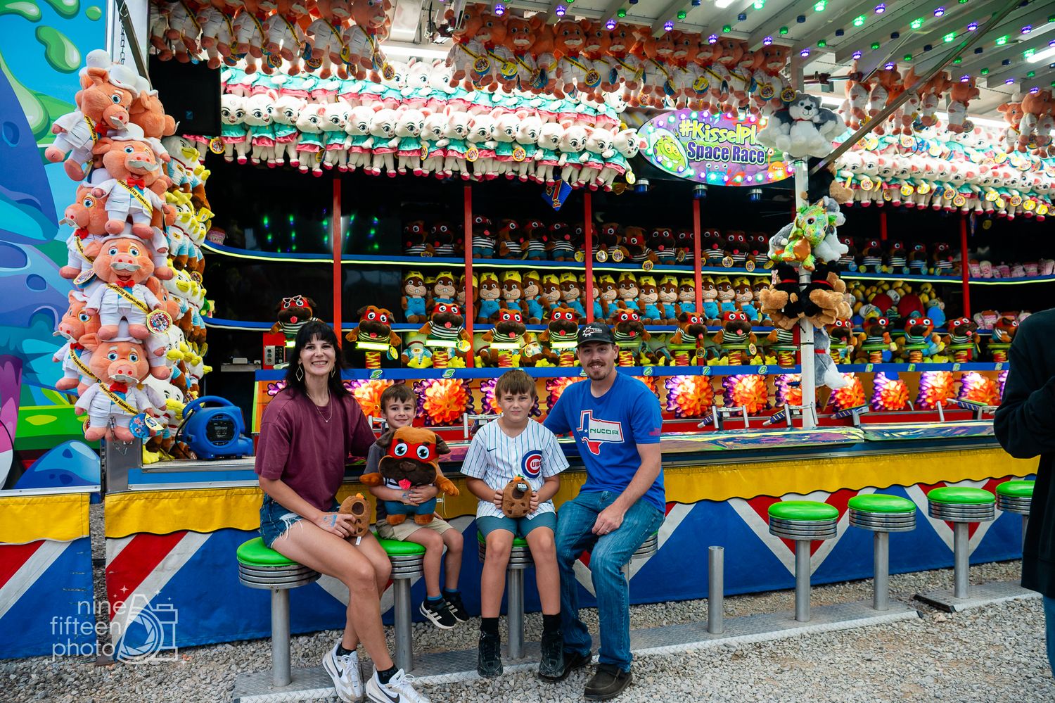 Family of four posing at a colorful carnival game booth filled with stuffed animal prizes at a fun fair.