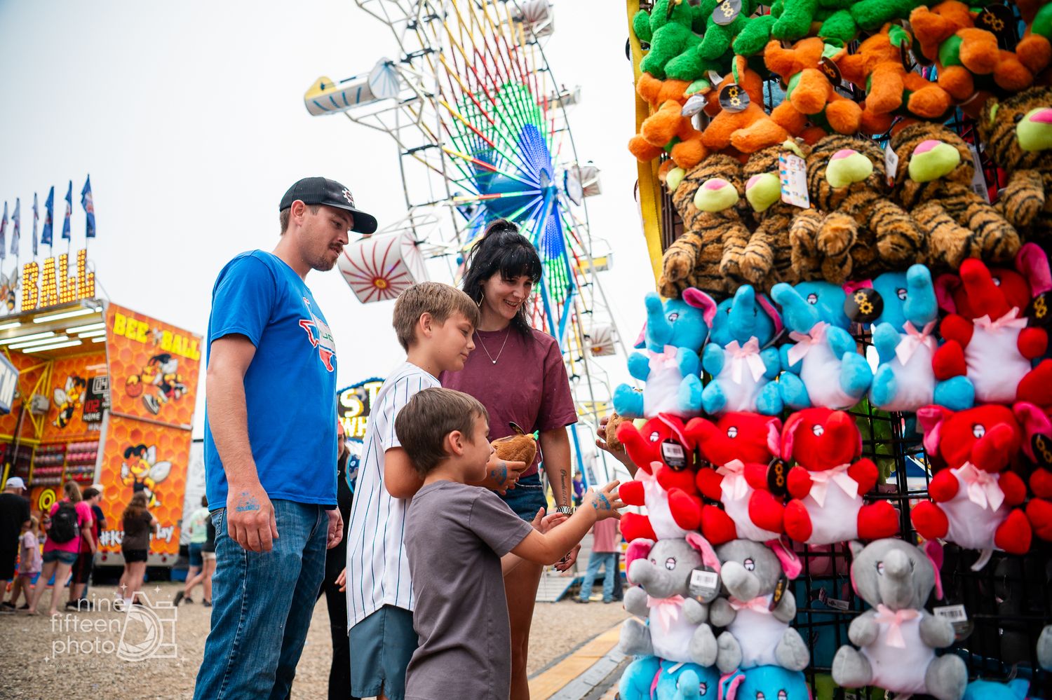 Family enjoying carnival games and colorful stuffed animal prizes at a fairground with a Ferris wheel in the background.