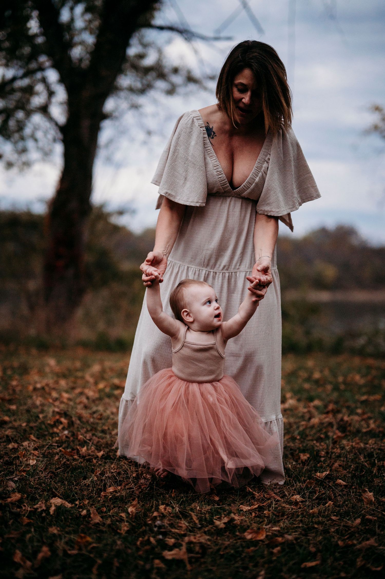 Mother in white dress helps toddler girl in pink tutu take first steps outdoors in autumn leaves.