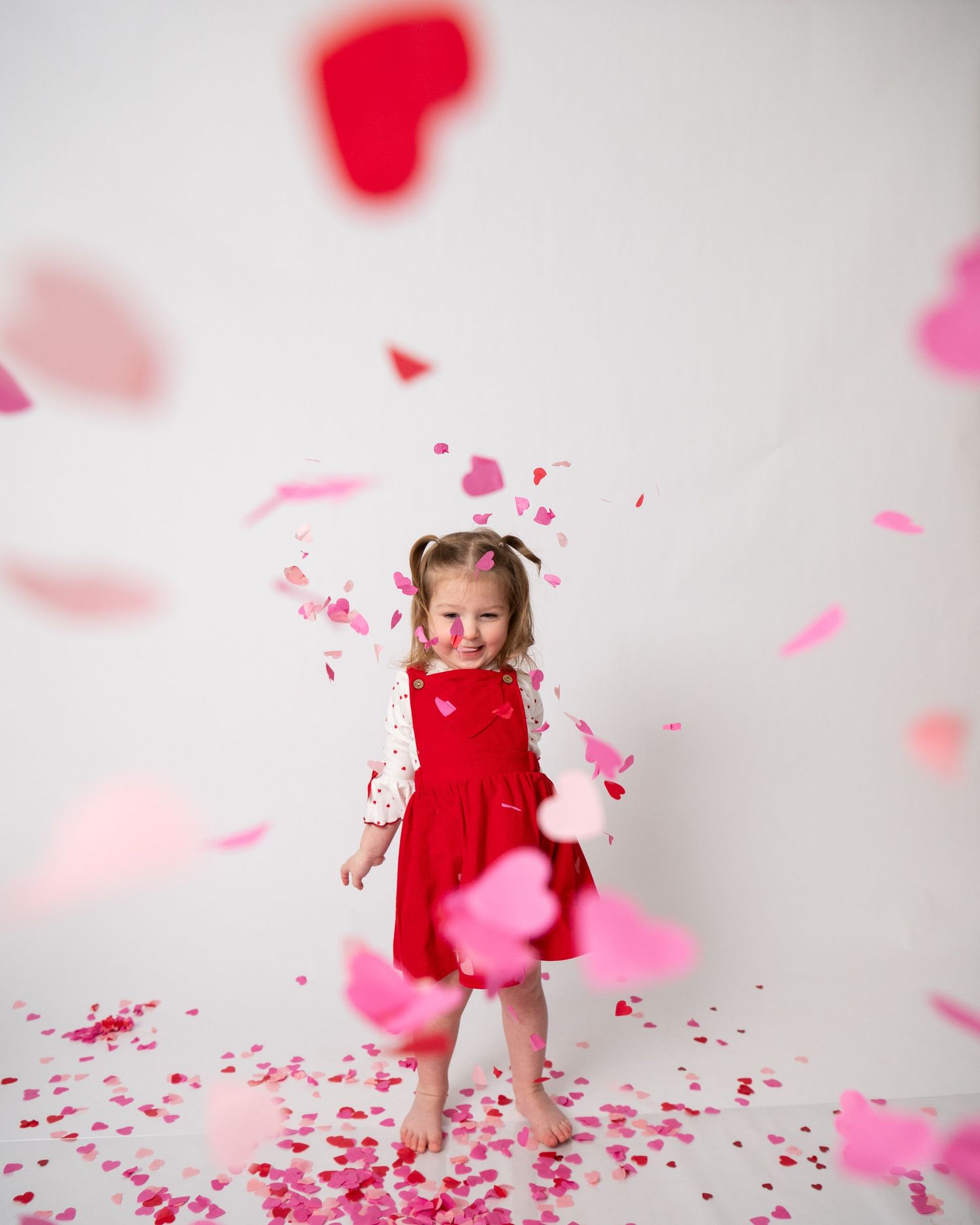 A joyful little girl in a red dress plays among falling pink and red heart confetti in a Valentine's Day photo shoot.