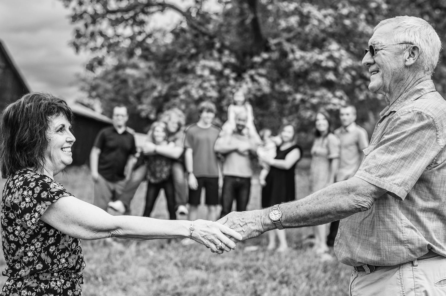 Black and white photo of couple dancing and holding hands while family cheers in background.