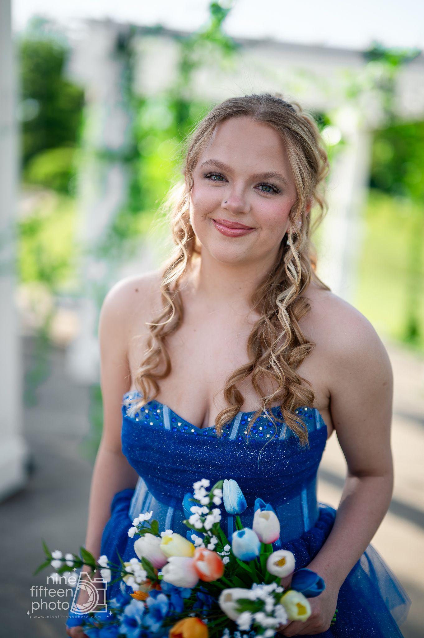 Young woman in blue strapless gown holding colorful tulip bouquet, smiling outdoors at formal event.