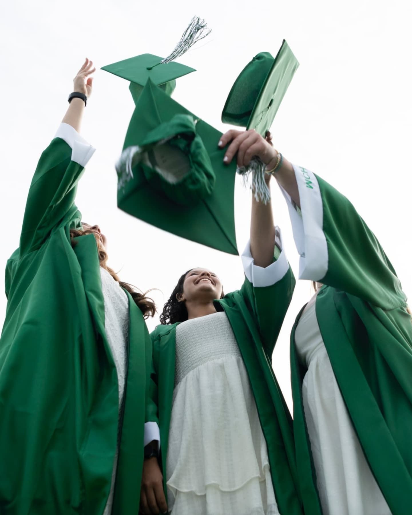 Three graduates in green caps and gowns joyfully toss their mortarboards into the sky.