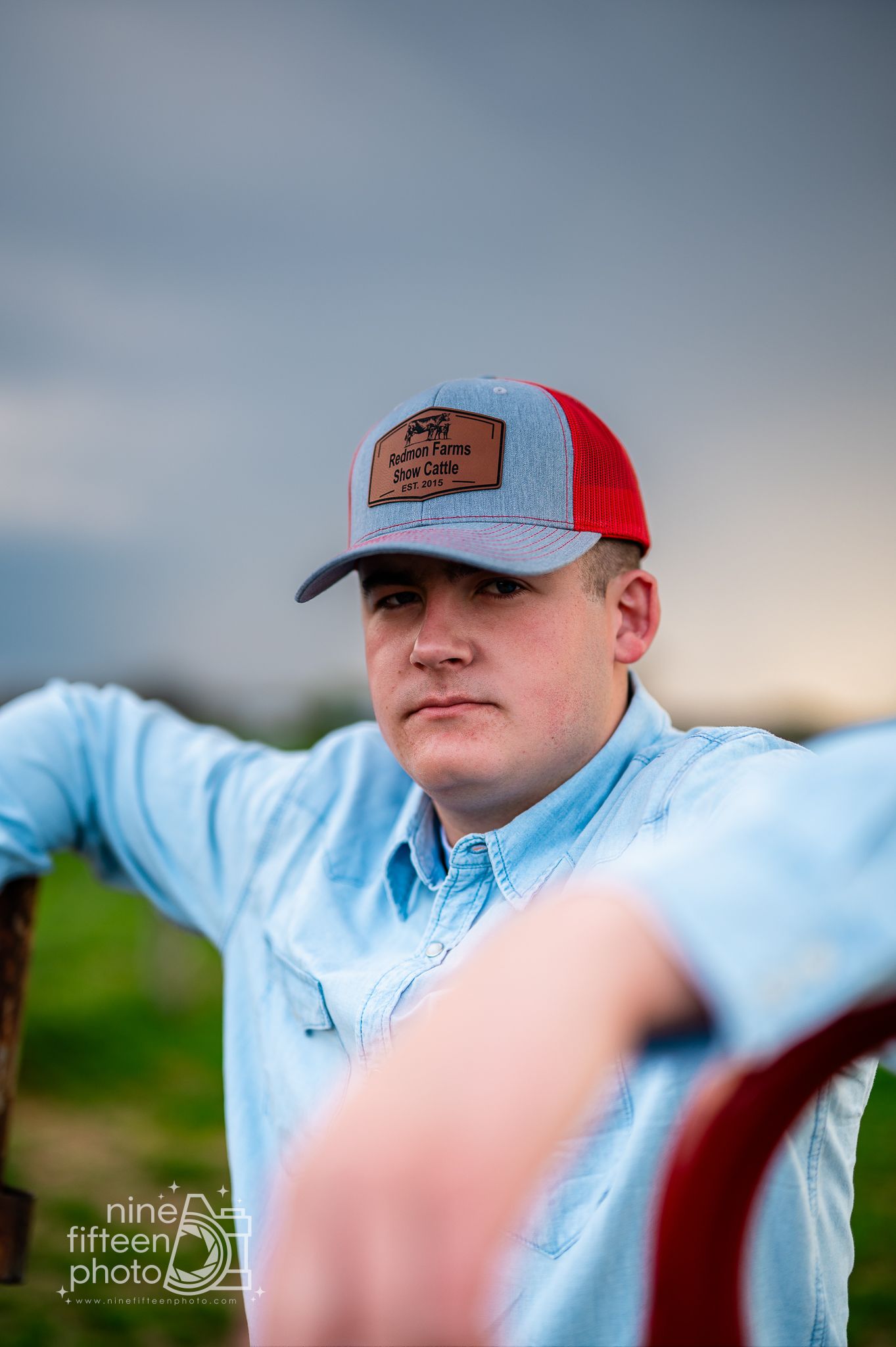 Young man in a denim shirt and red trucker hat poses outdoors against a dramatic cloudy sky.