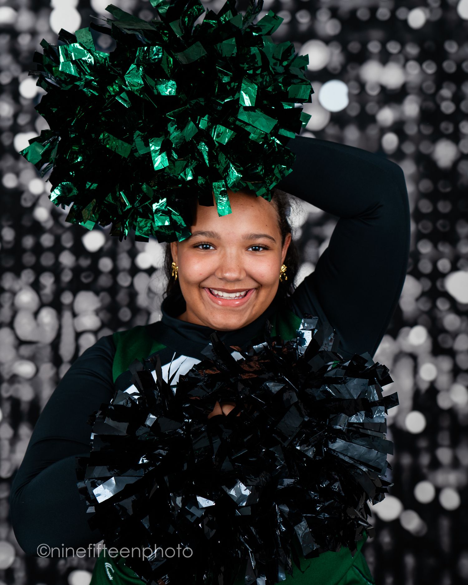 Smiling cheerleader holding green and black pom-poms against a silver bokeh backdrop in a dark uniform.