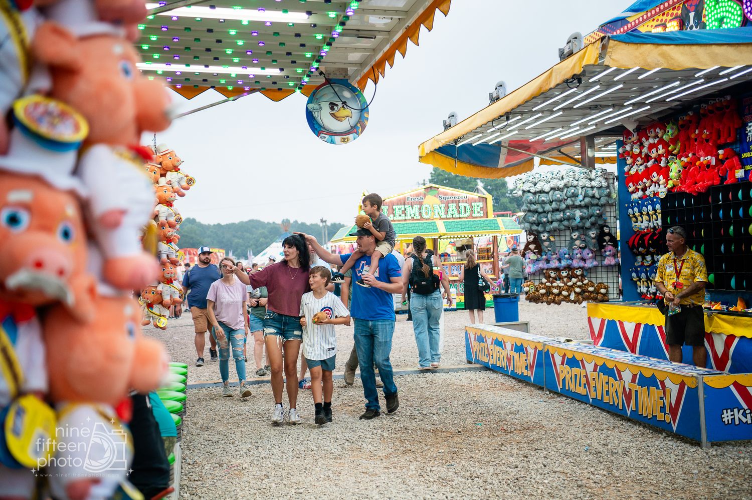 Families enjoy a lively carnival midway with colorful game booths, stuffed animal prizes, and lemonade stands at a summer fair.
