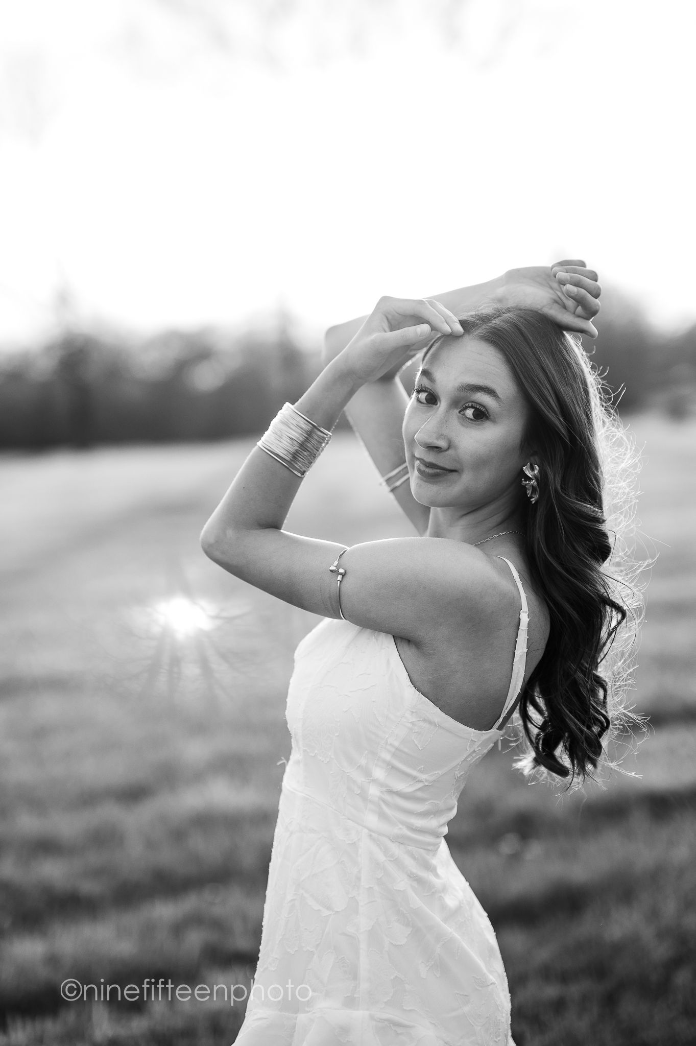 Black and white portrait of a young woman in a white dress posing gracefully in an open field at sunset.