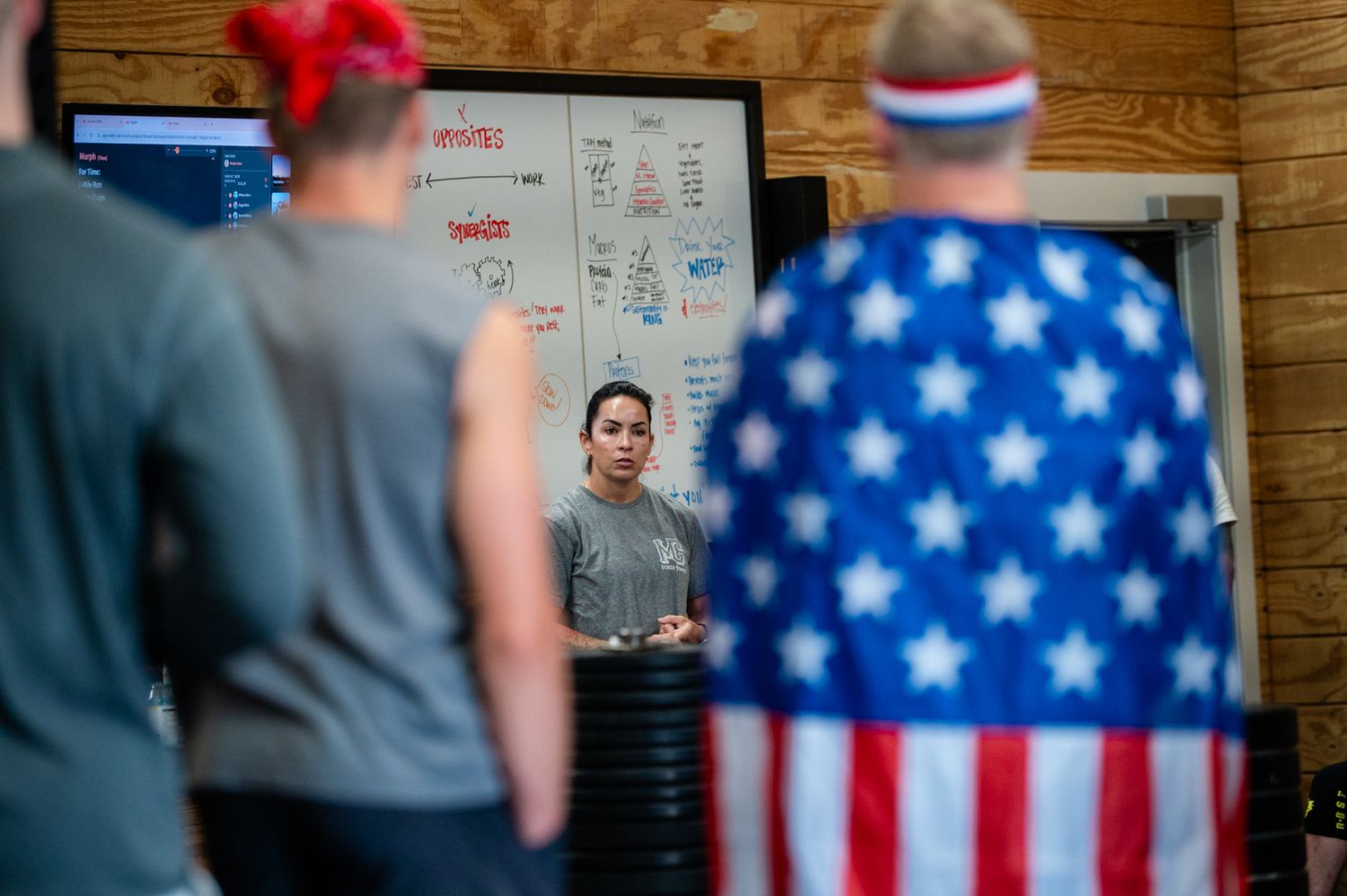 Woman coaching athletes in a gym, with American flag cape and MAGA hat visible in foreground.