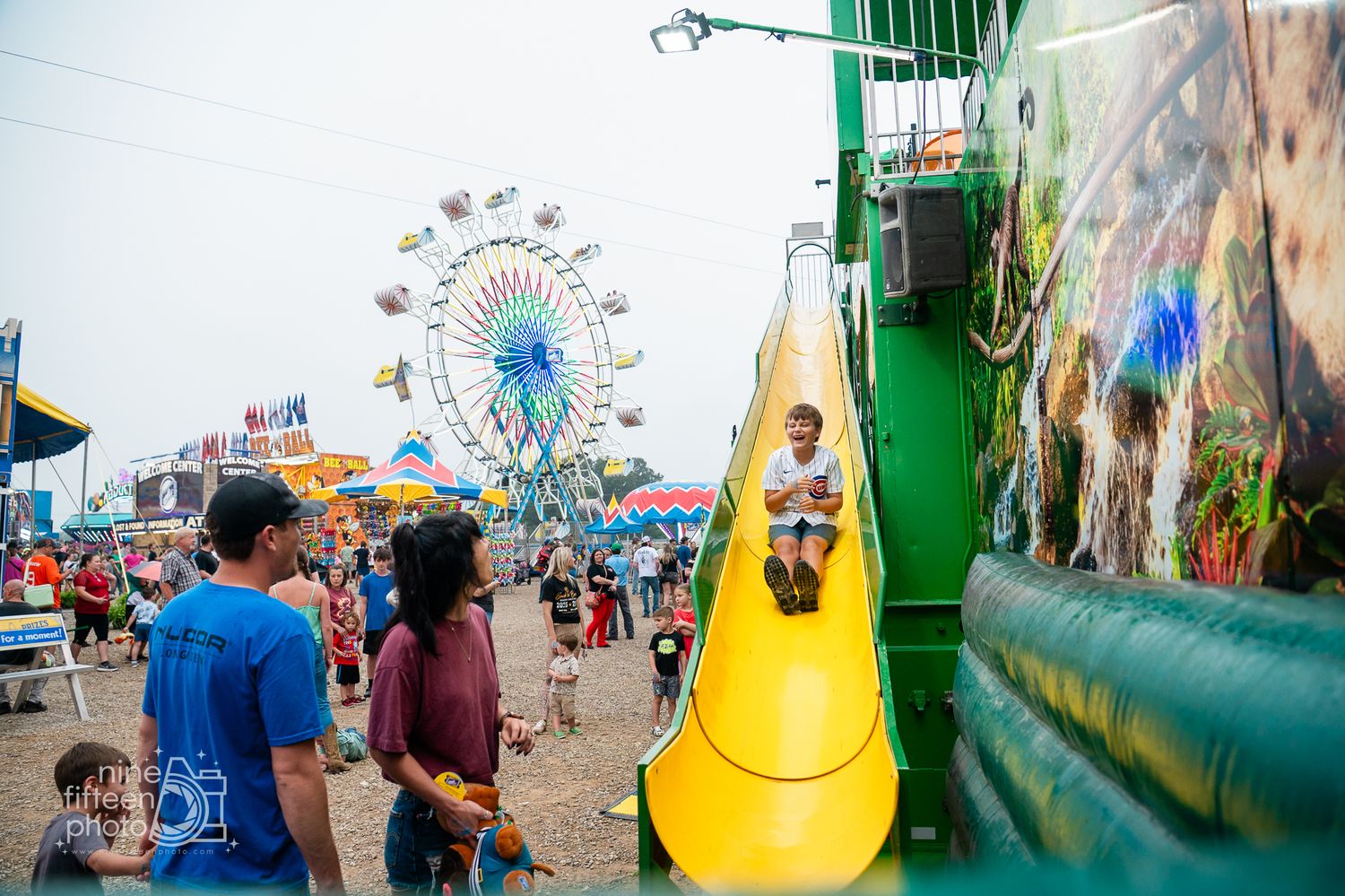 Child sliding down yellow slide at county fair with Ferris wheel in background.