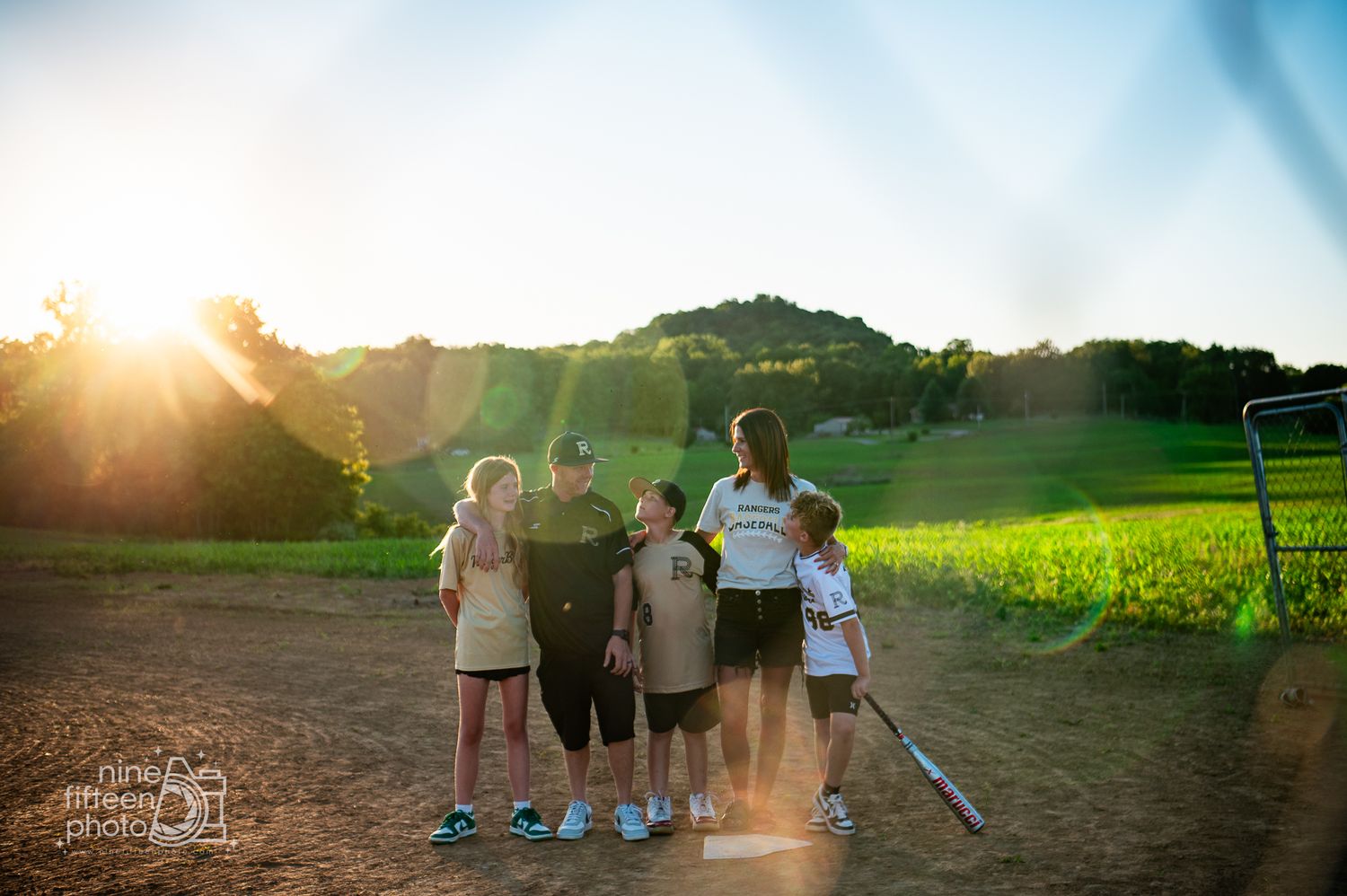 Family of five standing together on a dirt field at golden hour with lush green hills and sunburst in background.