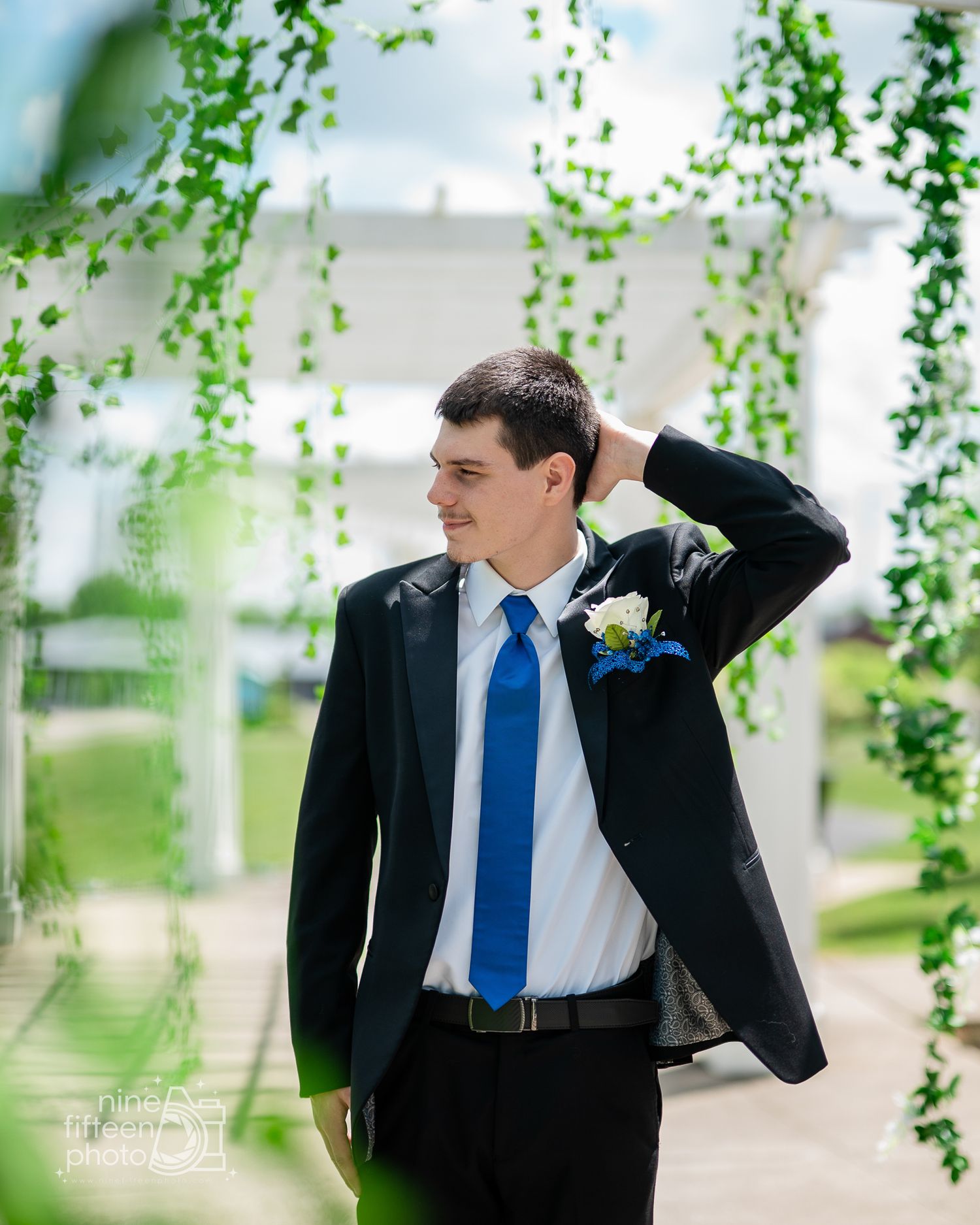 A groom in a black suit with blue tie and boutonniere poses outdoors under green vines at a wedding venue.