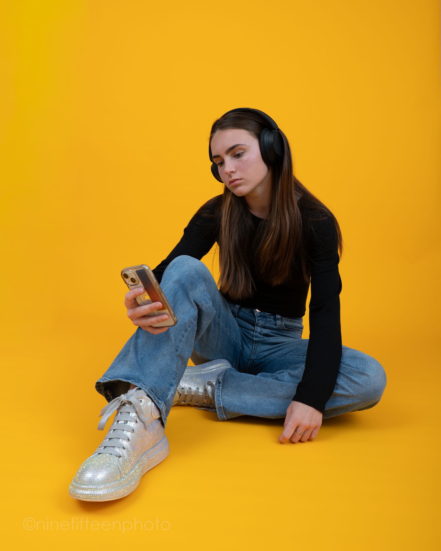 Teenage girl in black outfit sitting on yellow background wearing headphones and looking at her smartphone.