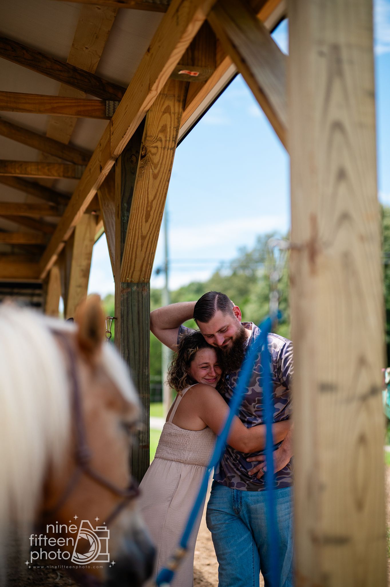 Couple embracing and smiling under a rustic wooden barn arch with a horse nearby on a sunny day.