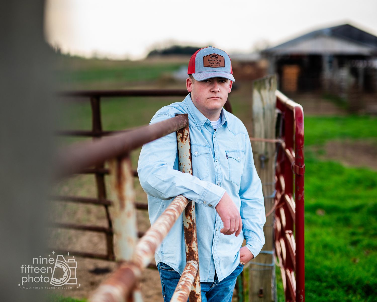 Teen boy in denim shirt and gray cap leans on rustic red farm gate during outdoor senior portrait session.