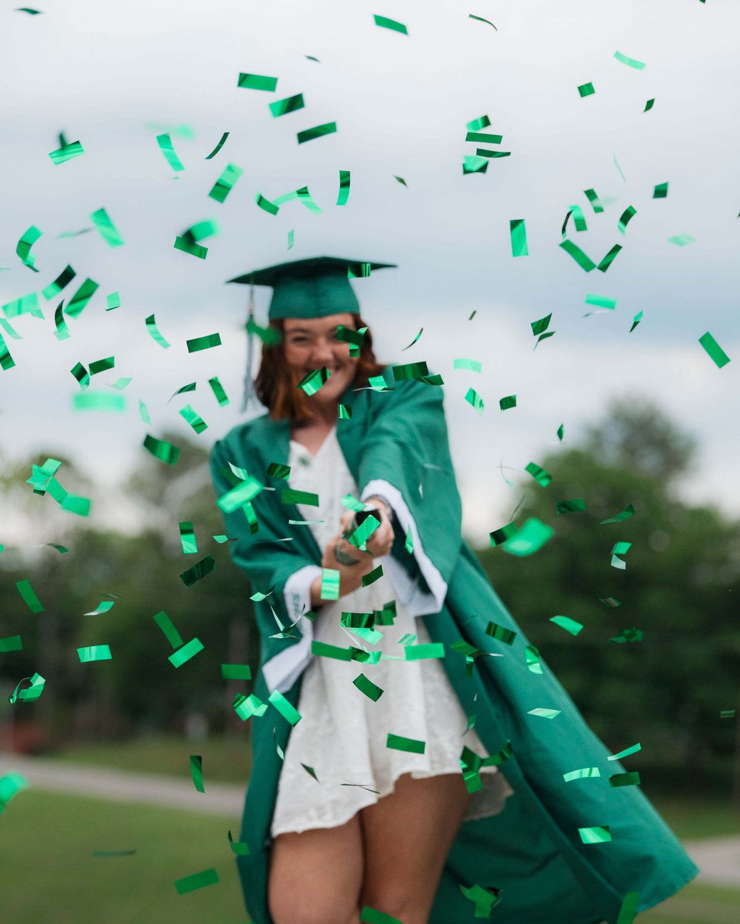 Smiling female graduate in green cap and gown celebrates surrounded by green confetti.