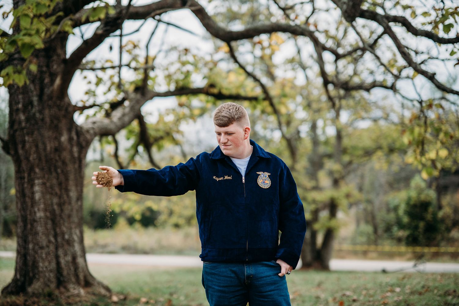 Young man in FFA jacket standing under a large oak tree holding an object in an outdoor park setting.