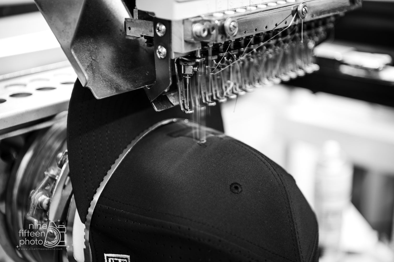 Close-up black and white photo of an embroidery machine stitching a black baseball cap.