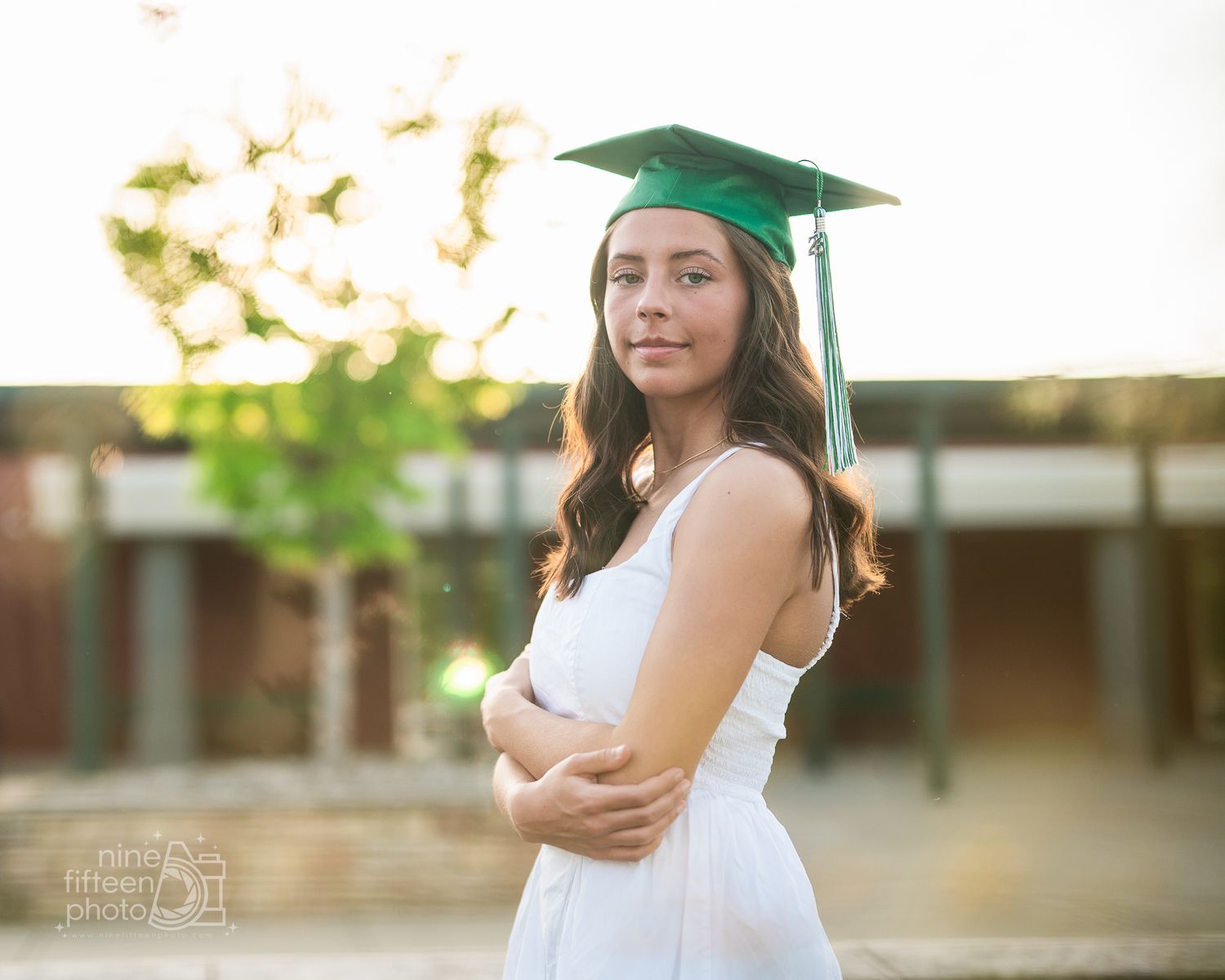 Young woman in white dress and green graduation cap poses outdoors during golden hour senior portrait session.