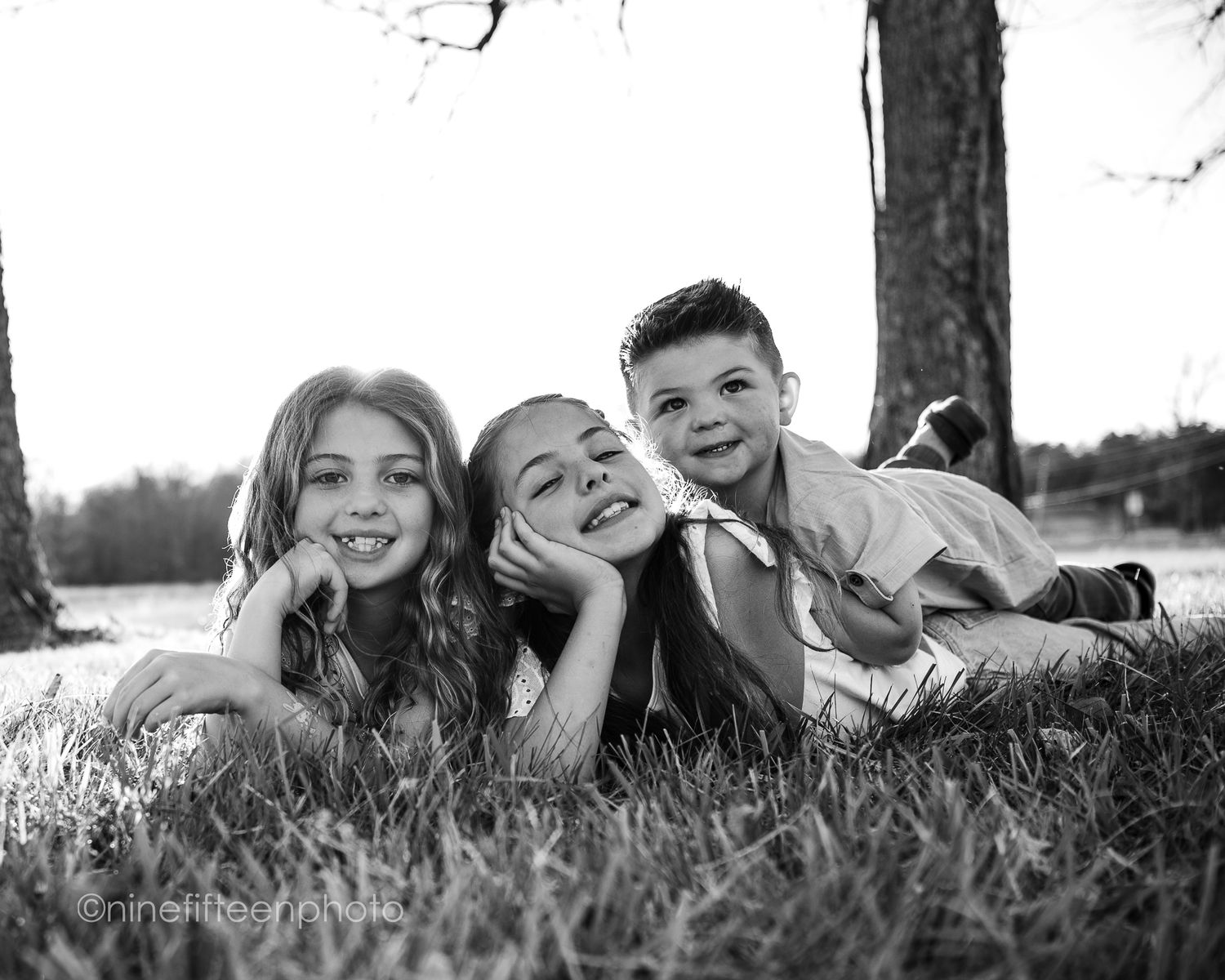 Three smiling children lying on grass in black and white portrait photo outdoors near trees.