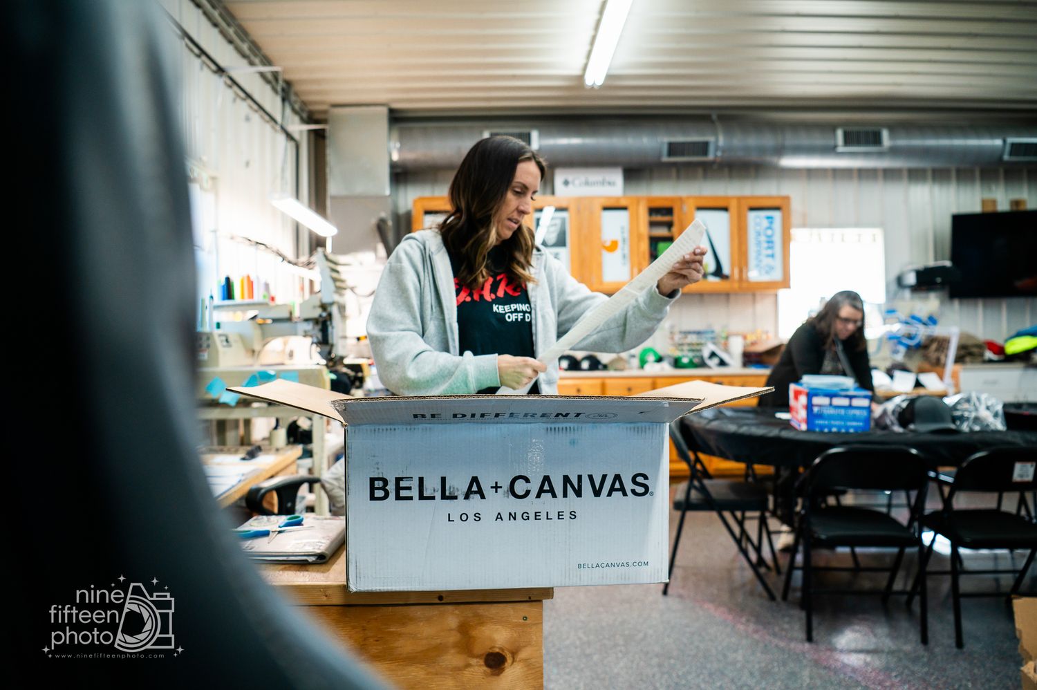 Young woman working at a Bella Canvas Los Angeles branded workstation in a busy workshop.