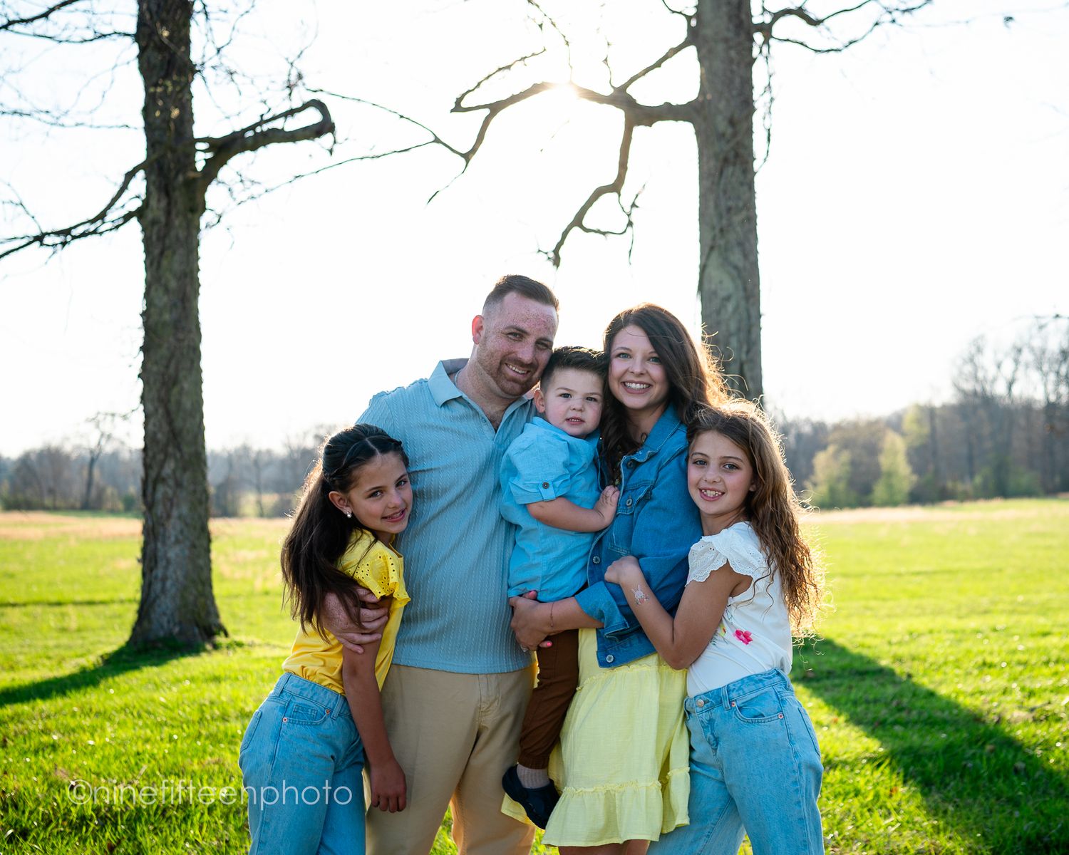 Happy family of five with three children smiling together in a sunny green park with bare trees behind them.
