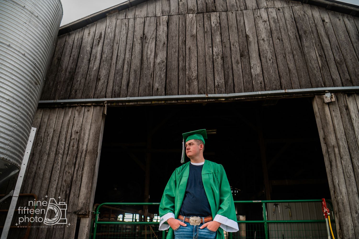 Graduate in green cap and gown stands proudly before a rustic wooden barn on a farm.