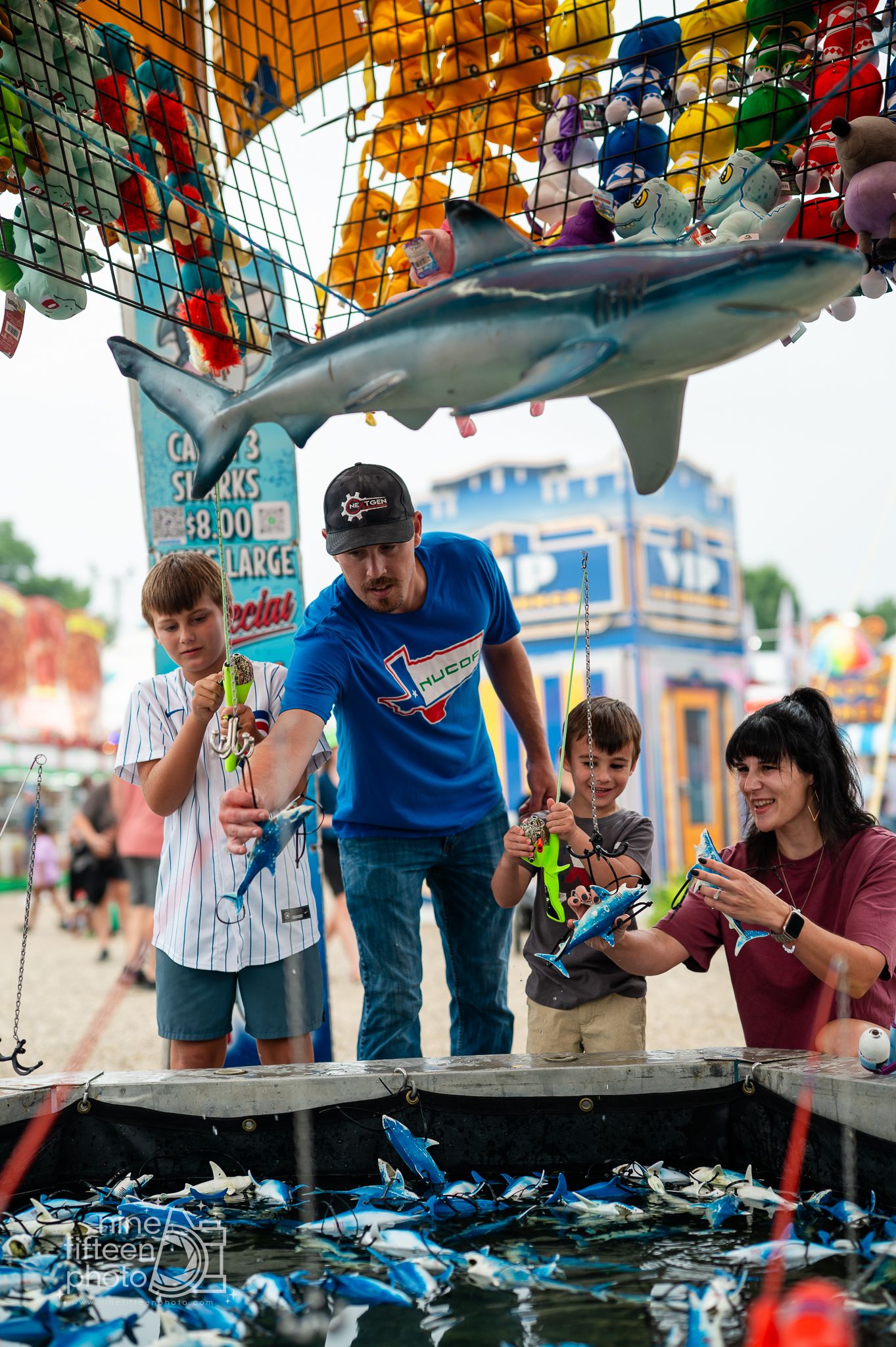 Family playing a carnival fishing game under a shark display at a colorful fair midway with prize stuffed animals.