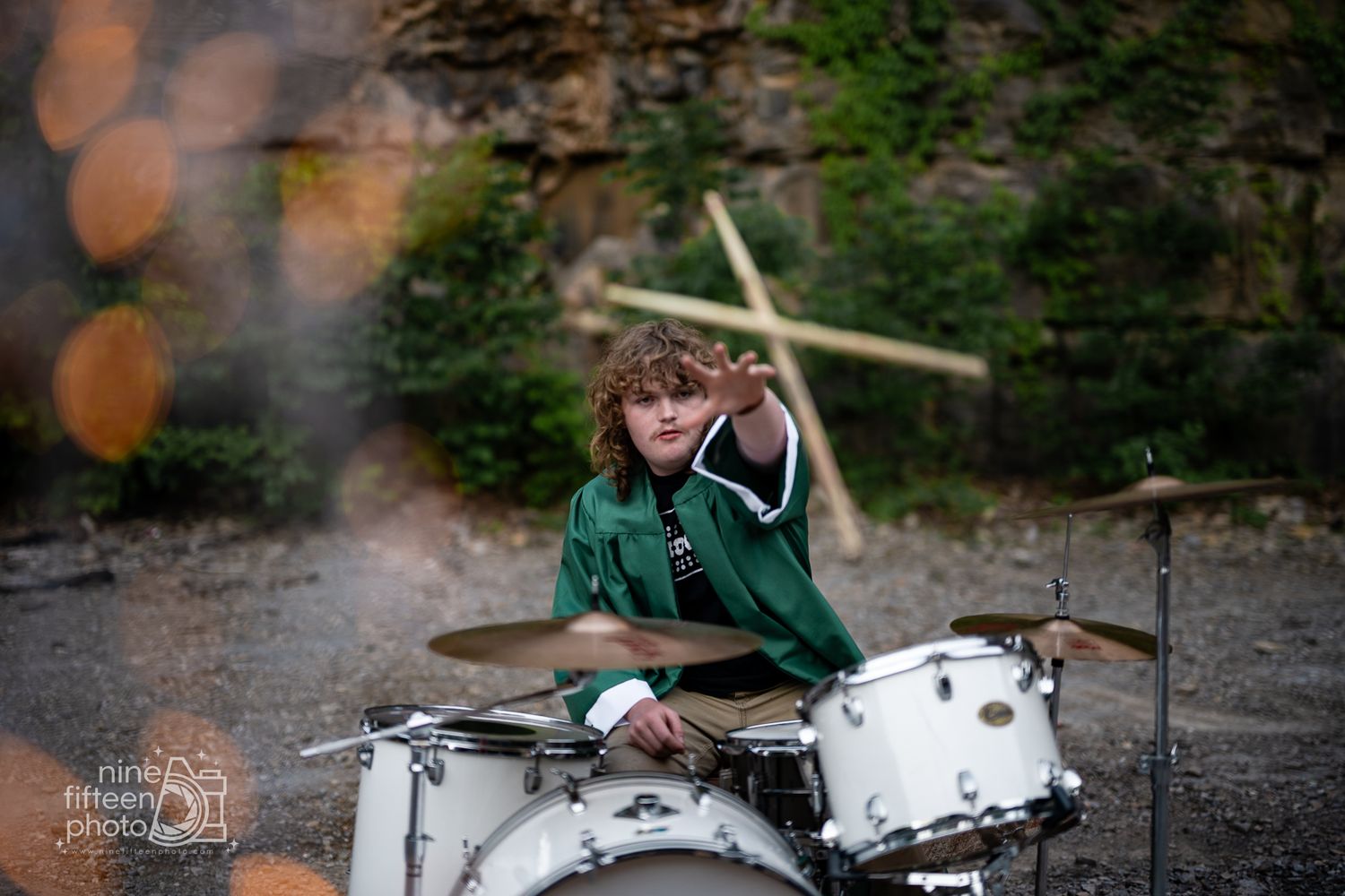 Young curly-haired drummer in green jacket plays white drum kit outdoors near rocky cliff, mid-performance.