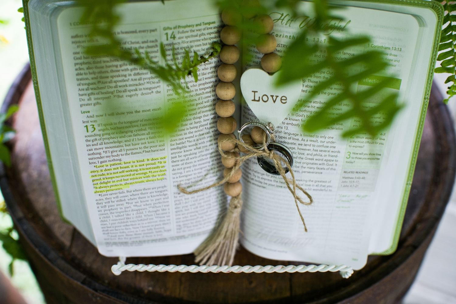 Open Bible with wooden bead garland and heart charm reading Love, displayed in rustic wooden barrel with greenery.