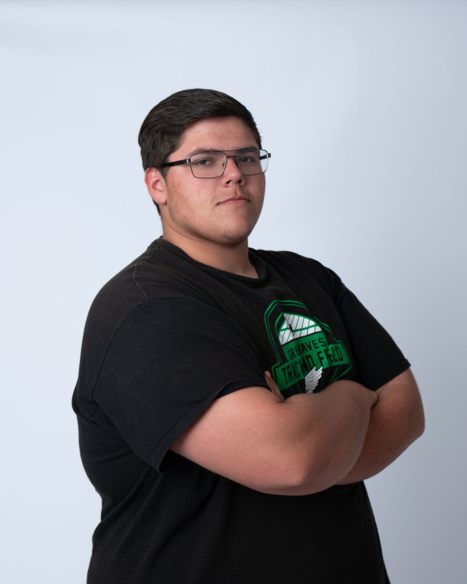 Young man in black graphic t-shirt with arms crossed poses against white background wearing glasses.