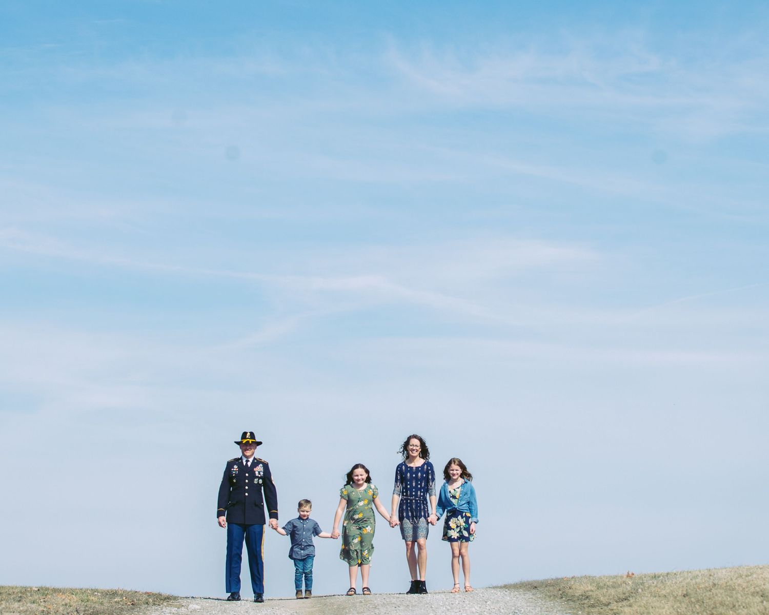 Military family of five standing together under wide open blue sky outdoors.