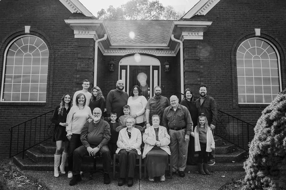 Large multigenerational family group portrait in black and white outside a historic brick building.