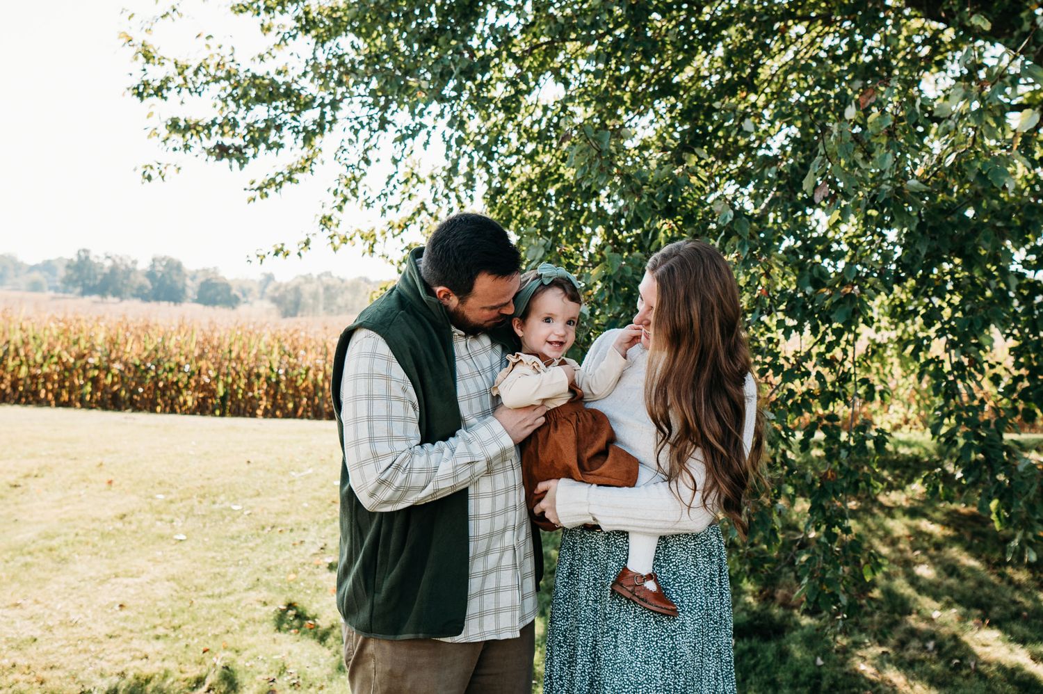Parents holding toddler under a tree sharing a sweet laugh during their fall family photo session.