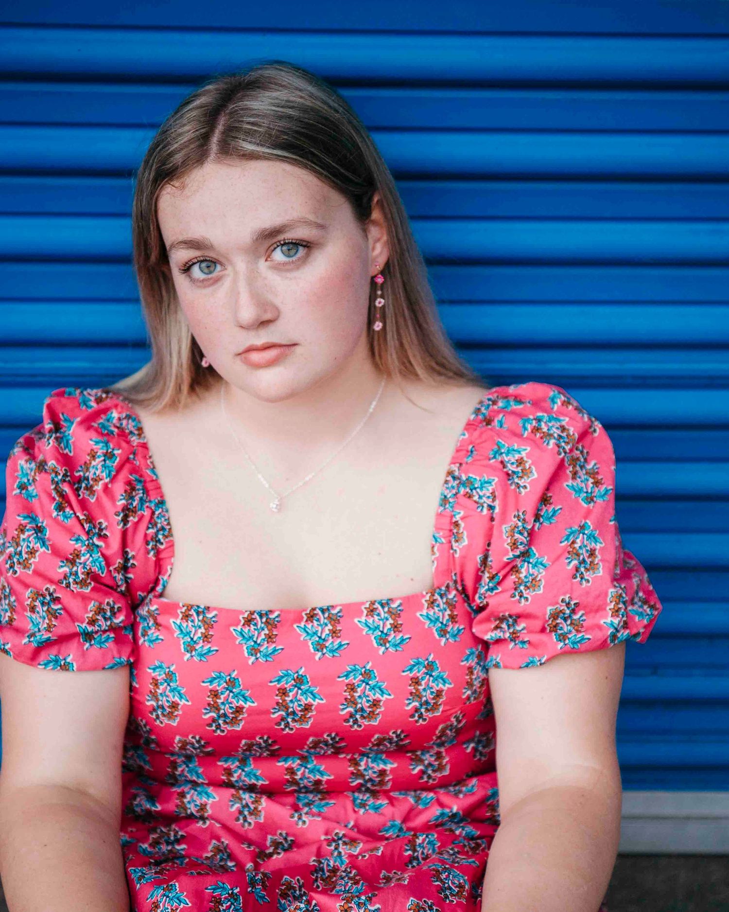 Young woman in red floral dress posing against a bright blue corrugated metal background.