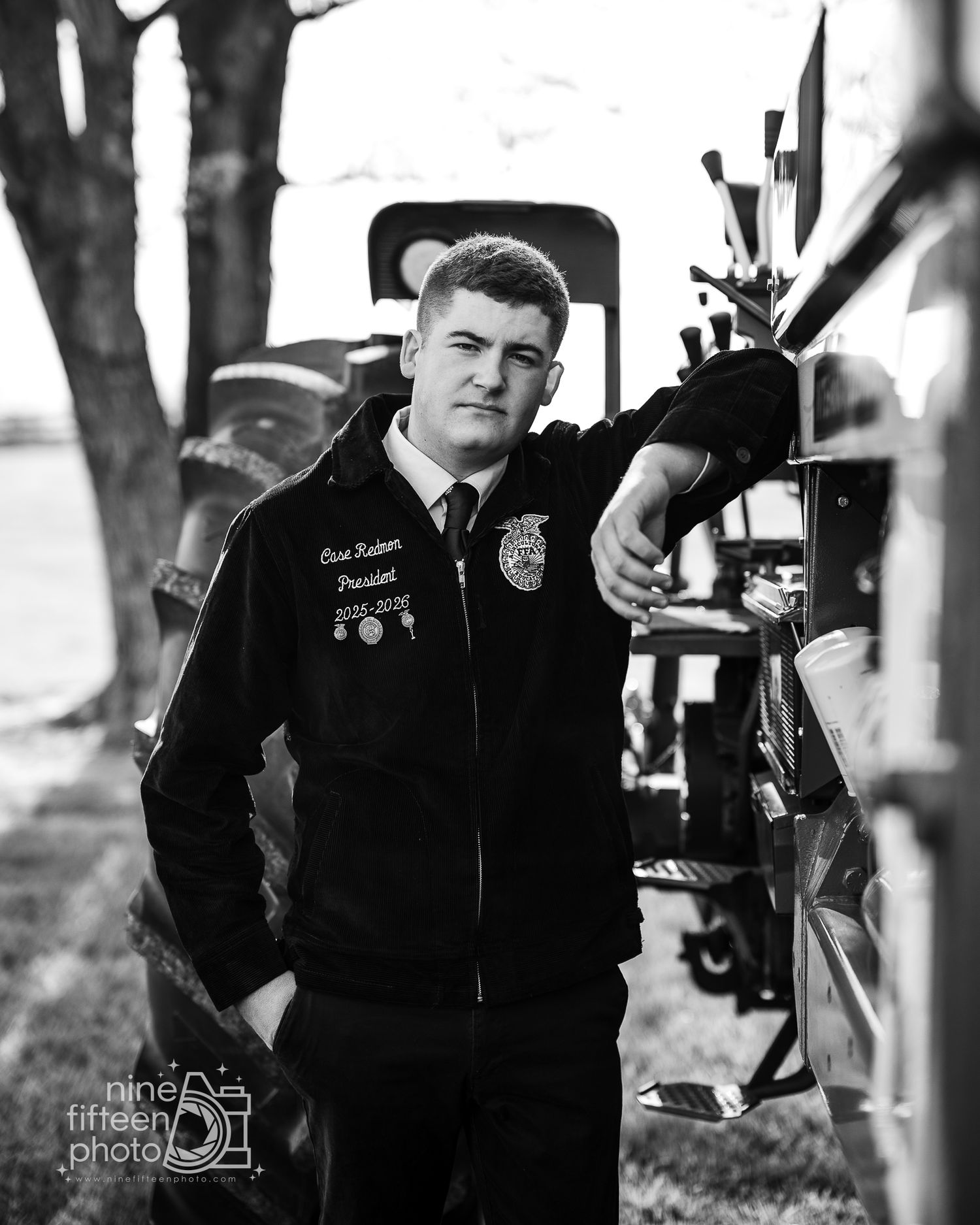 Black and white photo of a young man in an FFA jacket leaning against farm equipment outdoors.