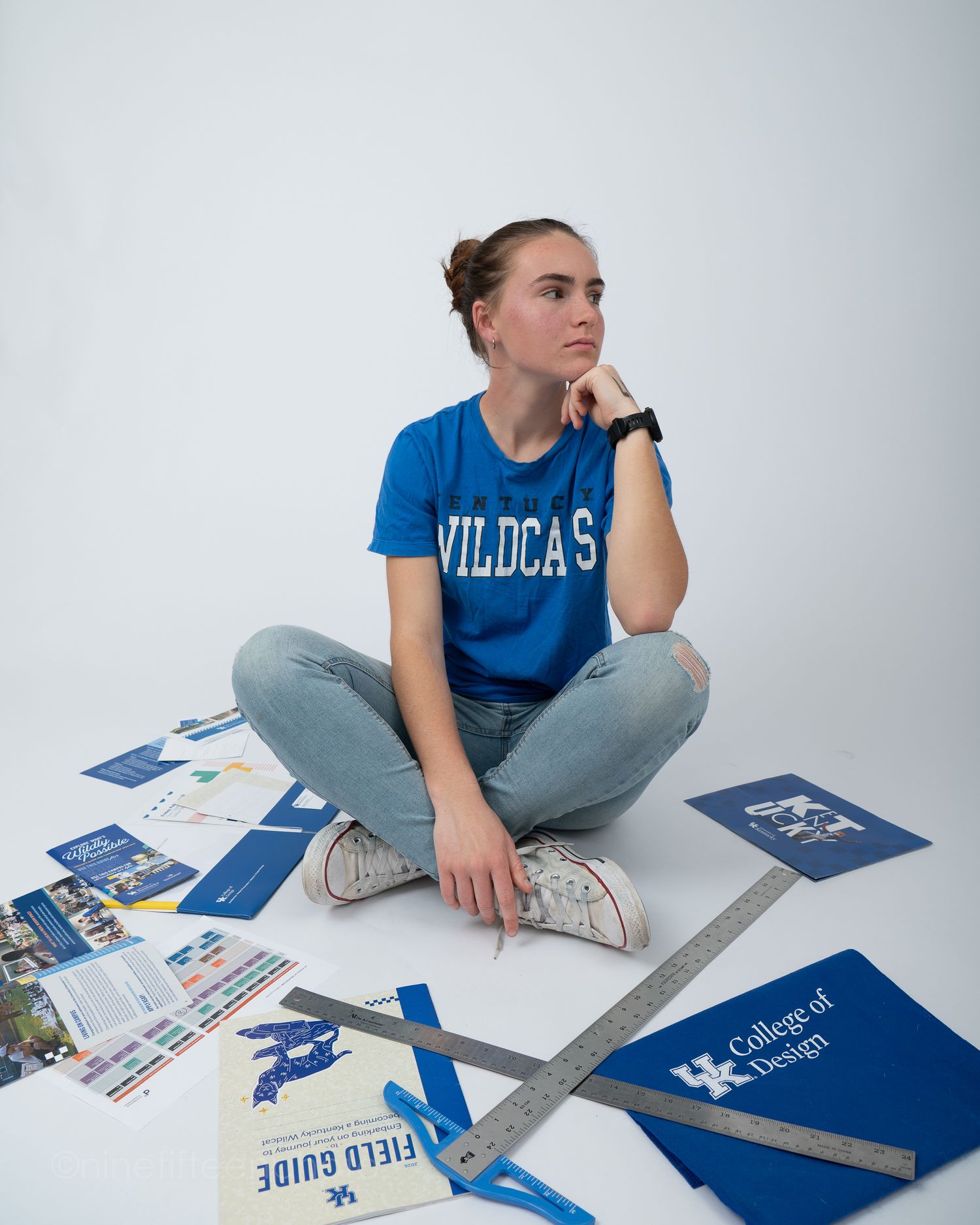 Female student in a blue Wildcats shirt sitting cross-legged surrounded by University of Kentucky College of Design materials.