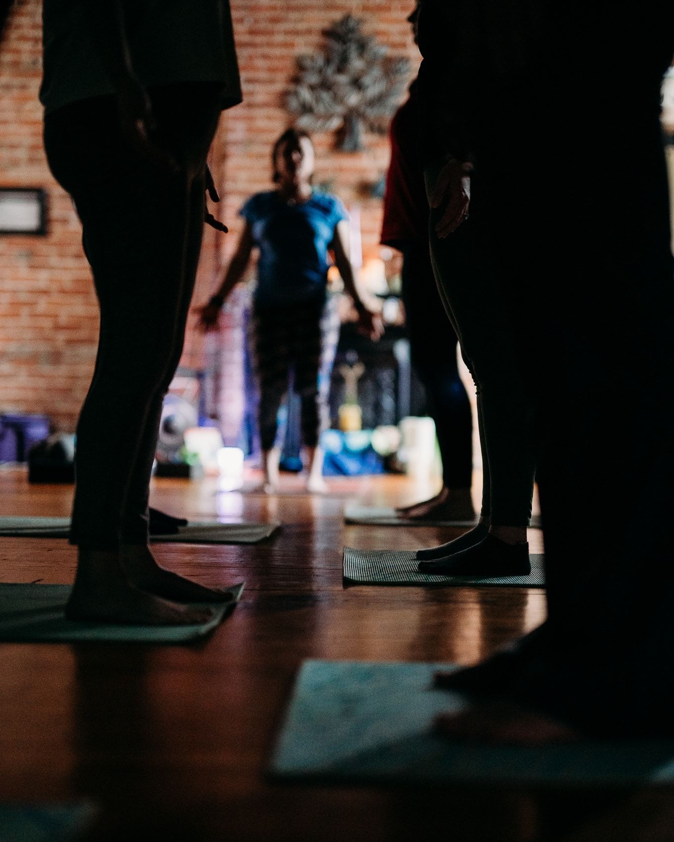 Silhouetted yoga students stand on mats in candlelit brick-walled studio with instructor in background.